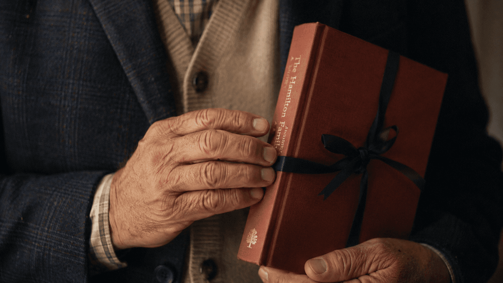 An older person's weathered hands holding a new terracotta hardcover book tied with a thin grosgrain ribbon